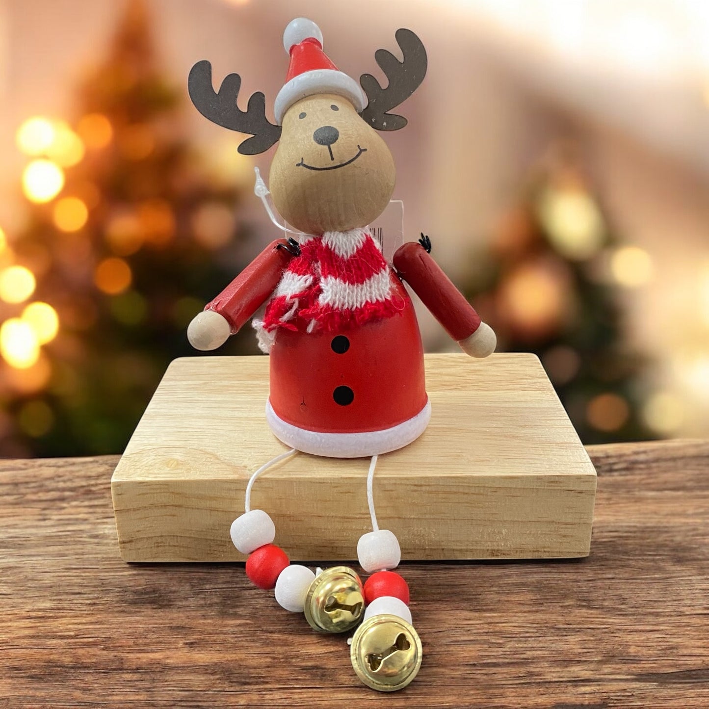Holzfigur eines Elchs in roter Kleidung mit Schal und goldenen Glöckchen, sitzt auf einem Holzblock vor weihnachtlicher Beleuchtung.
