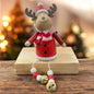 Holzfigur eines Elchs in roter Kleidung mit Schal und goldenen Glöckchen, sitzt auf einem Holzblock vor weihnachtlicher Beleuchtung.
