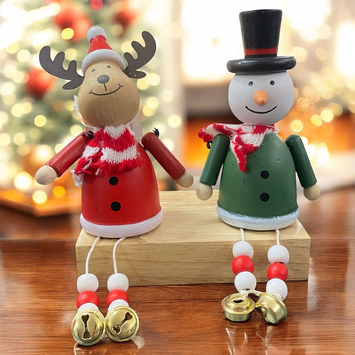 Zwei weihnachtliche Holzfiguren, ein Elch in roter Kleidung und ein Schneemann in grüner Kleidung, beide mit Schal und goldenen Glöckchen an den Beinen, sitzen auf einem Holzblock im Ambiente