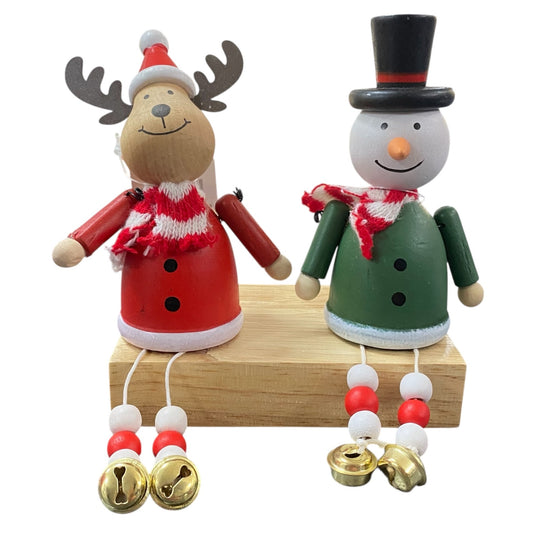 Zwei weihnachtliche Holzfiguren, ein Elch in roter Kleidung und ein Schneemann in grüner Kleidung, beide mit Schal und goldenen Glöckchen an den Beinen, sitzen auf einem Holzblock.