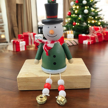 Holzfigur eines Schneemanns mit grünem Mantel, rotem Schal und schwarzem Zylinder, sitzt auf einem Holzblock vor einem geschmückten Weihnachtsbaum.

