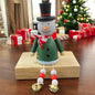 Holzfigur eines Schneemanns mit grünem Mantel, rotem Schal und schwarzem Zylinder, sitzt auf einem Holzblock vor einem geschmückten Weihnachtsbaum.

