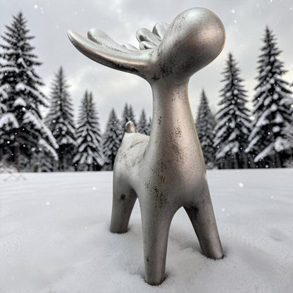 Silberfarbene Dekofigur in Form eines stilisierten Elchs im Schnee vor einem Hintergrund mit verschneiten Bäumen.