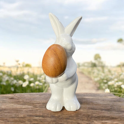 Ein weißer Osterhase aus weißer Keramik, der ein Holz Osterei in der Hand trägt im Ambiente