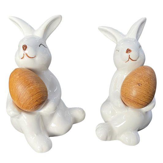 Zwei creme-weiße Deko Osterhasen mit aus Keramik, sitzend mit braunem Holzei im Arm vor weißem Hintergrund