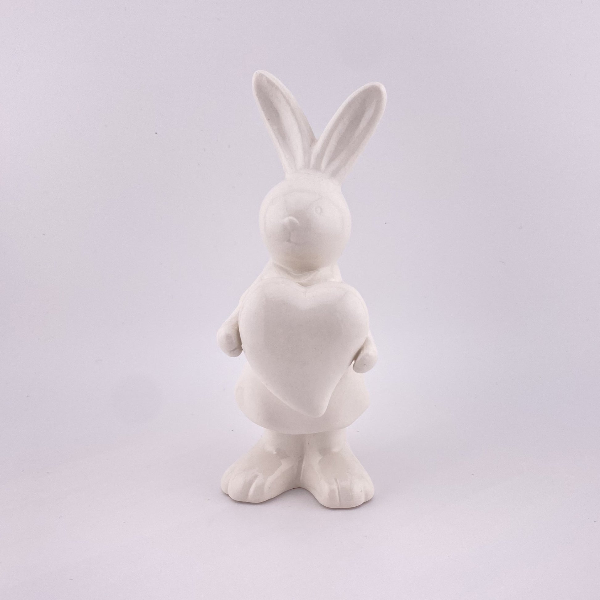  Ein weiblicher Deko Osterhase aus creme-weißer Keramik, der die Ohren gespitzt und ein Herz in ihren Pfoten hält. Sie ist vor weißem Hintergrund platziert.