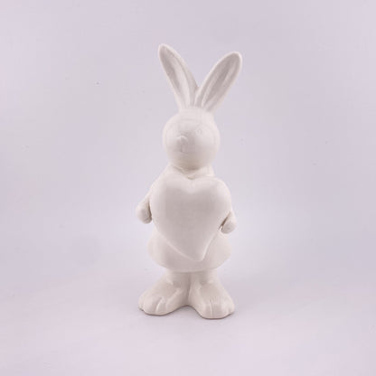  Ein weiblicher Deko Osterhase aus creme-weißer Keramik, der die Ohren gespitzt und ein Herz in ihren Pfoten hält. Sie ist vor weißem Hintergrund platziert.