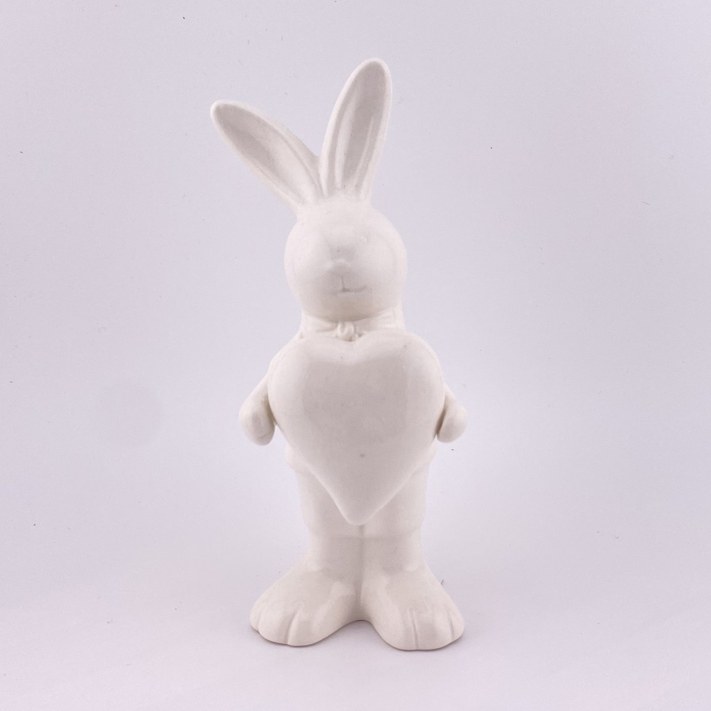 Ein männlicher Deko Osterhase aus creme-weißer Keramik, der die Ohren gespitzt und ein Herz in ihren Pfoten hält. Sie ist vor weißem Hintergrund platziert.