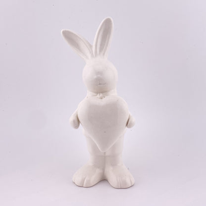 Ein männlicher Deko Osterhase aus creme-weißer Keramik, der die Ohren gespitzt und ein Herz in ihren Pfoten hält. Sie ist vor weißem Hintergrund platziert.