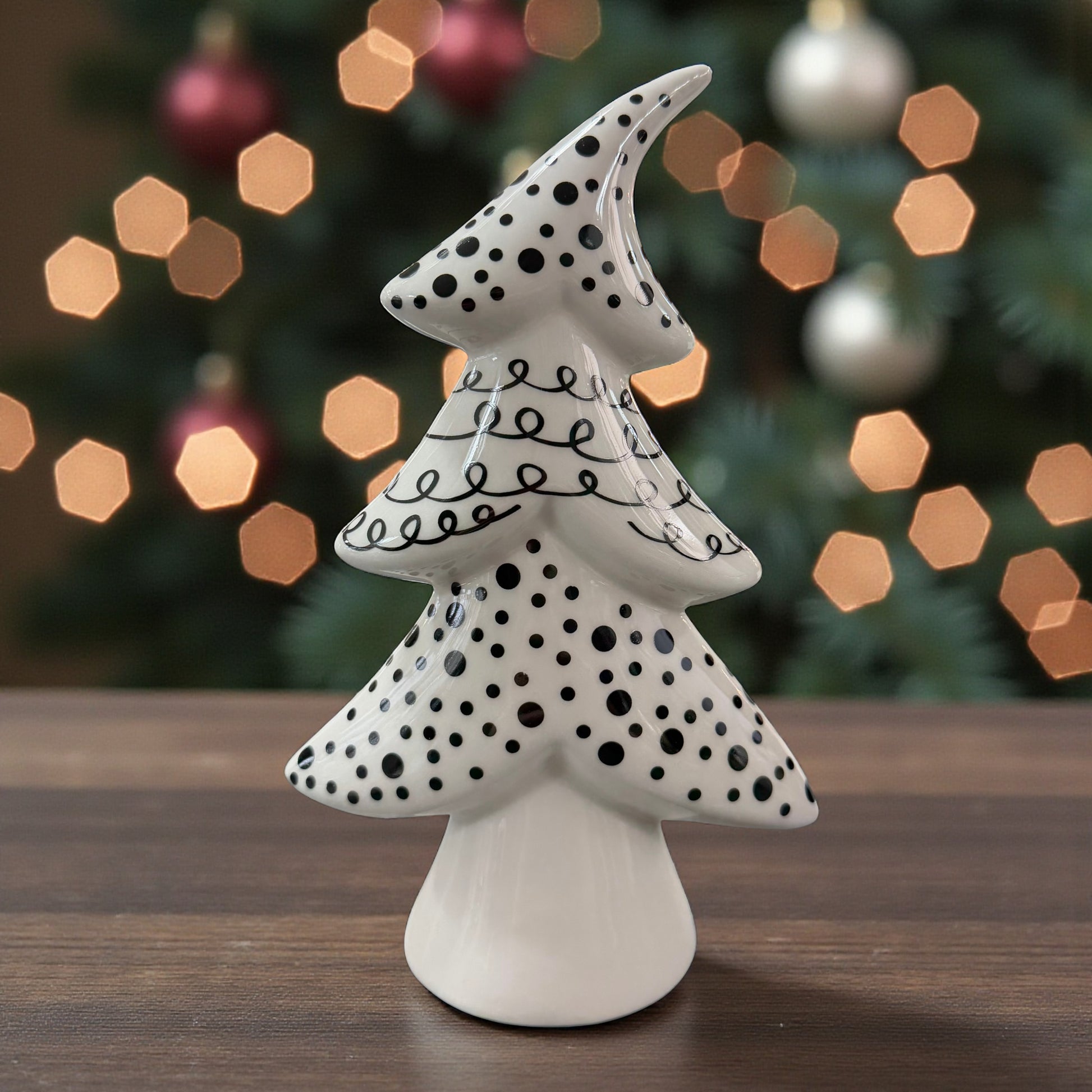 Ein weißer Keramik-Tannenbaum mit schwarzen Punkten und geschwungenen Linien steht auf einem Tisch vor einem weihnachtlich beleuchteten Hintergrund.