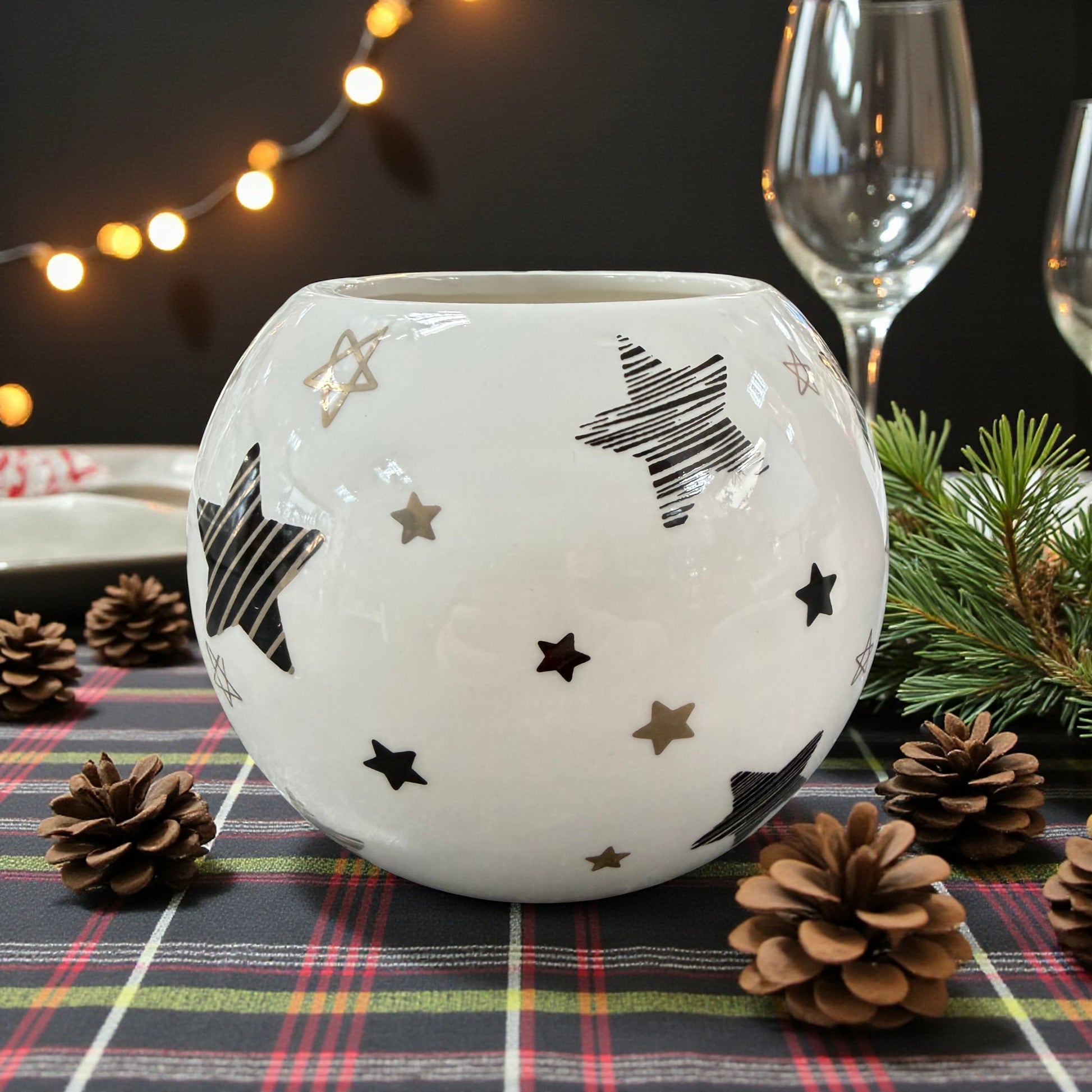 Kugelvase aus weißer Keramik mit dekorativen Sternen in Schwarz und Gold, platziert auf kariertem Tischläufer mit Tannenzweigen und Zapfen, vor weihnachtlicher Tischdekoration mit Lichterkette und Gläsern im Hintergrund.
