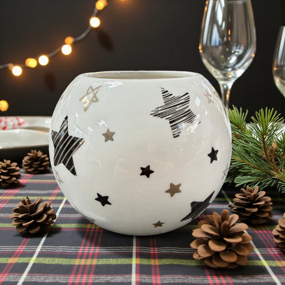 Kugelvase aus weißer Keramik mit dekorativen Sternen in Schwarz und Gold, platziert auf kariertem Tischläufer mit Tannenzweigen und Zapfen, vor weihnachtlicher Tischdekoration mit Lichterkette und Gläsern im Hintergrund.