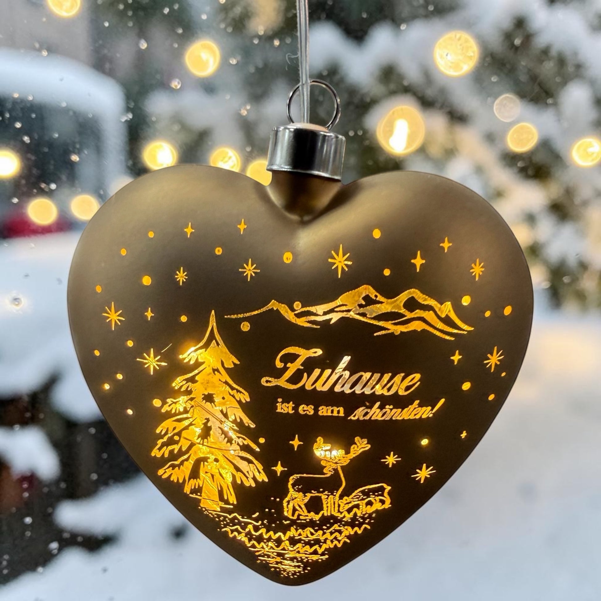 Ein herzförmiger, brauner LED-Dekoanhänger mit warmweißem Licht zeigt eine winterliche Szene mit Tannenbaum, Hirsch und dem Schriftzug „Zuhause ist es am schönsten!“ vor verschneitem Hintergrund.