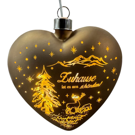 Ein herzförmiger, brauner LED-Dekoanhänger mit warmweißem Licht zeigt eine winterliche Szene mit Tannenbaum, Hirsch und dem Schriftzug „Zuhause ist es am schönsten!“.