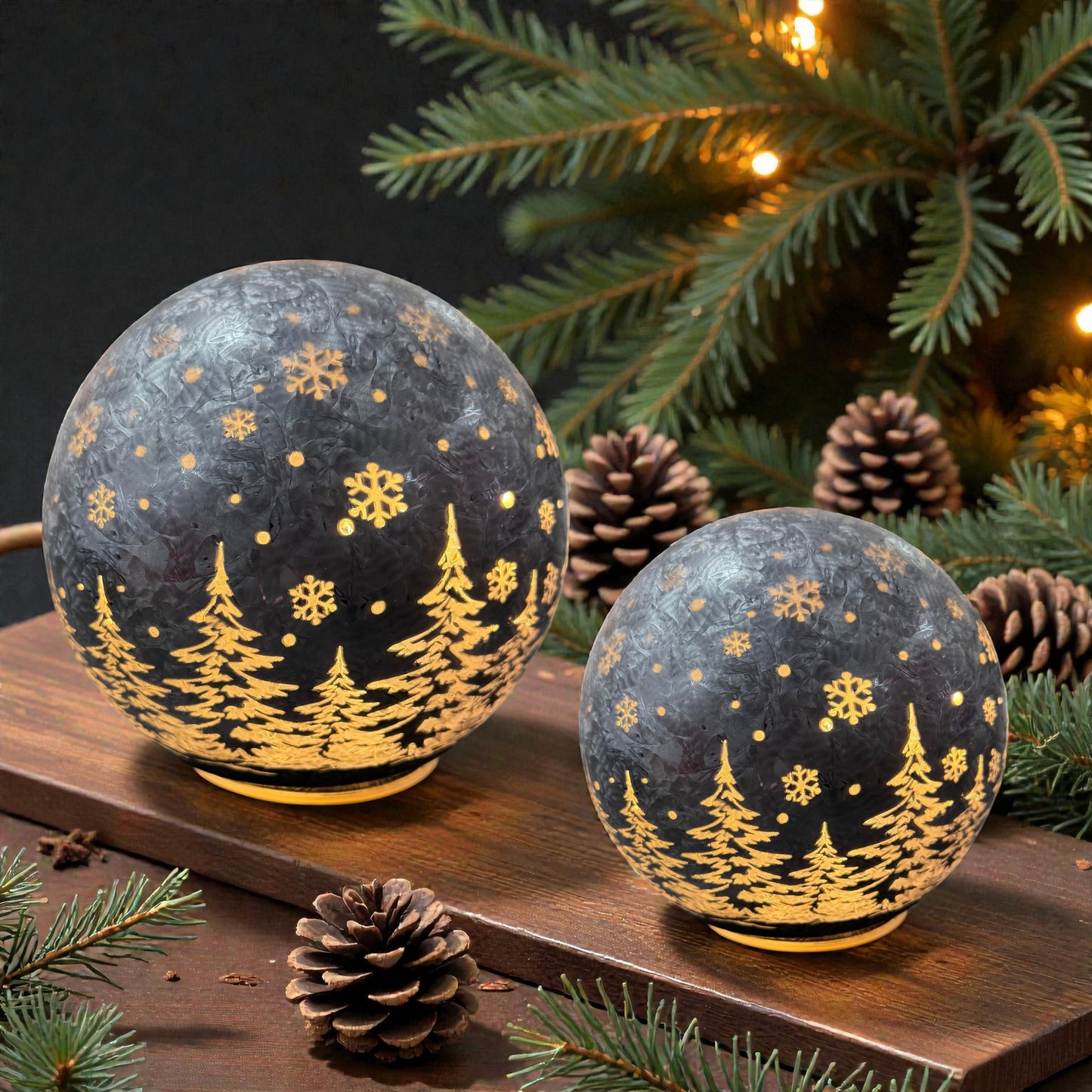 Zwei leuchtende LED-Dekokugeln in Anthrazit mit winterlichem Tannenbaum- und Schneeflockenmotiv stehen auf einer Holzoberfläche neben Tannenzweigen und Tannenzapfen.