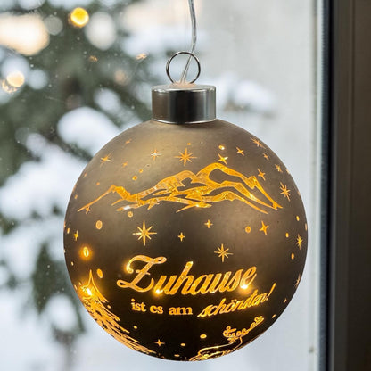 Eine runde, braune LED-Dekokugel zum Aufhängen mit warmweißem Licht zeigt eine winterliche Szene mit Bergen, Tannenbaum, Hirsch und dem Schriftzug „Zuhause ist es am schönsten!“ vor verschneitem Hintergrund.