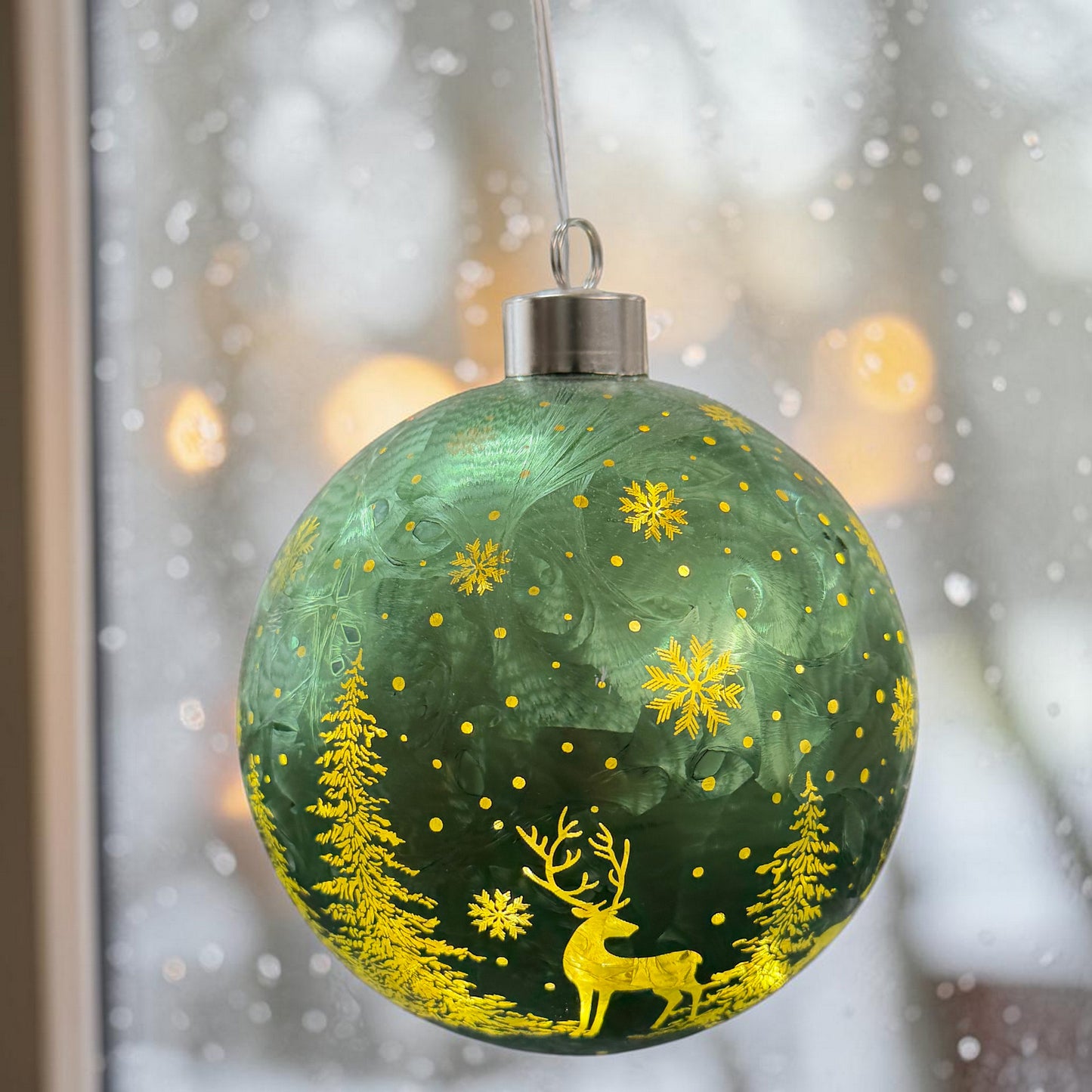 Grüne Kugeldekoration mit Hirsch-, Tannenbaum- und Schneeflockenmuster vor winterlichem Hintergrund.