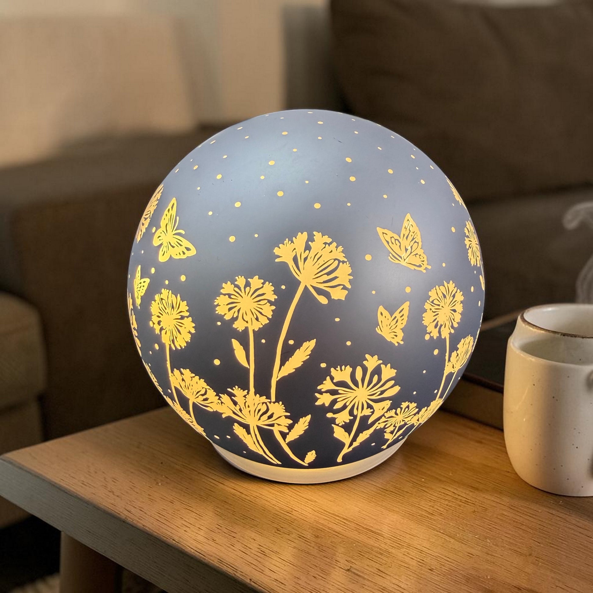 Leuchtende Kugellampe mit Blumen- und Schmetterlingsmotiven steht auf einem Holztisch neben einer Tasse.