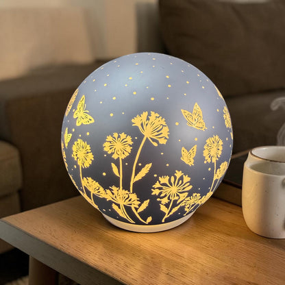 Leuchtende Kugellampe mit Blumen- und Schmetterlingsmotiven steht auf einem Holztisch neben einer Tasse.