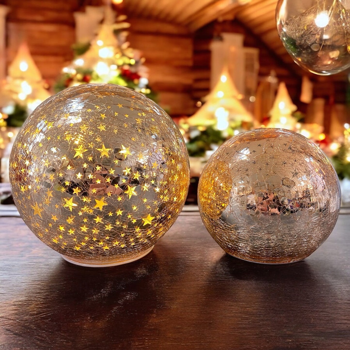 Zwei leuchtende rosa Crackle-Glas-Kugeln mit goldenen Sternen auf einem Holztisch vor warmem Lichterhintergrund.