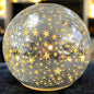 Leuchtende silberne Crackle-Glas-Kugel mit goldenen Sternenmotiven, strahlend im warmen Licht eines weihnachtlichen Ambientes.