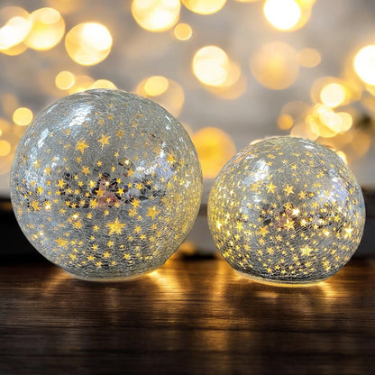 Zwei leuchtende silberne Crackle-Glas-Kugeln mit goldenen Sternen auf einem Holztisch vor warmem Lichterhintergrund.