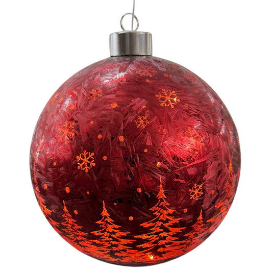 Rot leuchtende Kugeldekoration mit Tannenbaum- und Schneeflockenmuster vor verschneiter Fensterkulisse.