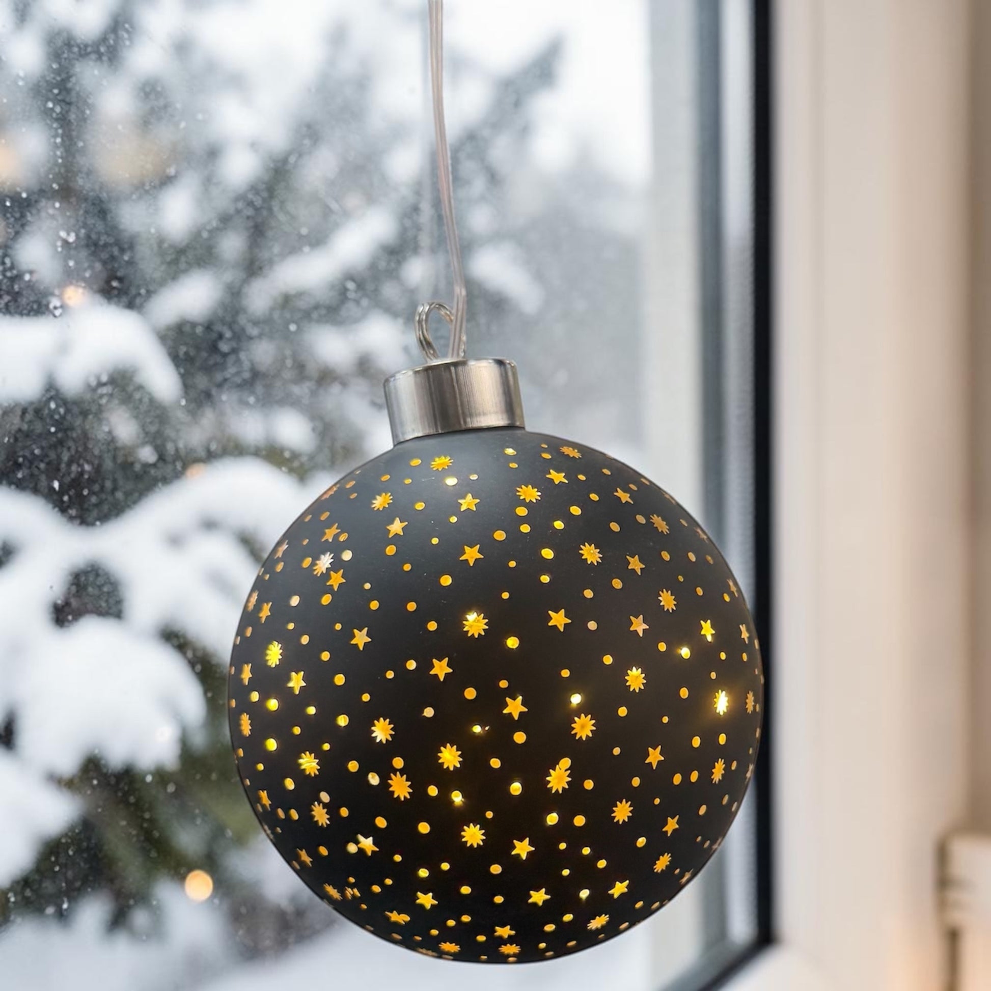 Eine schwarze LED-Dekokugel mit Sternenmuster und leuchtenden Punkten ist vor einem Fenster mit winterlichem Hintergrund platziert.