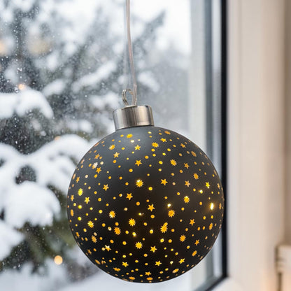 Eine schwarze LED-Dekokugel mit Sternenmuster und leuchtenden Punkten ist vor einem Fenster mit winterlichem Hintergrund platziert.