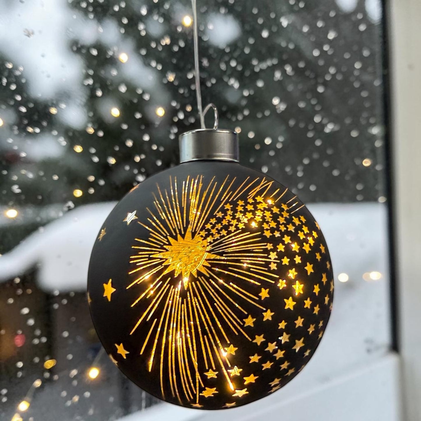 Eine schwarze LED-Dekokugel mit Sternschnuppen- und Sternmuster hängt vor einer winterlichen Außenszene.