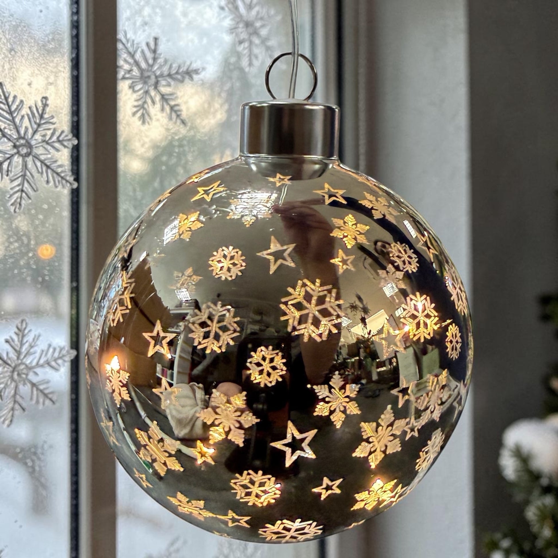 Eine glänzende silberne LED-Dekokugel mit ausgestanzten Mustern hängt vor einem Fenster mit sichtbaren Schneeflocken.