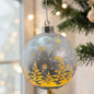 Graue Kugeldekoration mit goldfarbenen Tannenbäumen und Schneeflocken hängt vor unscharfem Lichterhintergrund.
