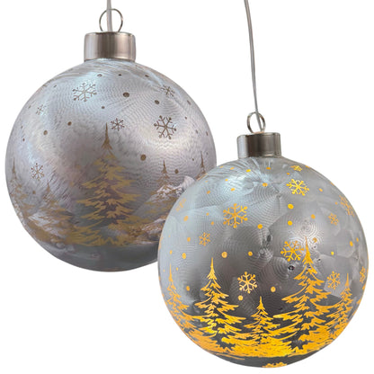 Zwei silberfarbene Kugeldekorationen mit Winterwaldmotiv und warm leuchtenden Details.