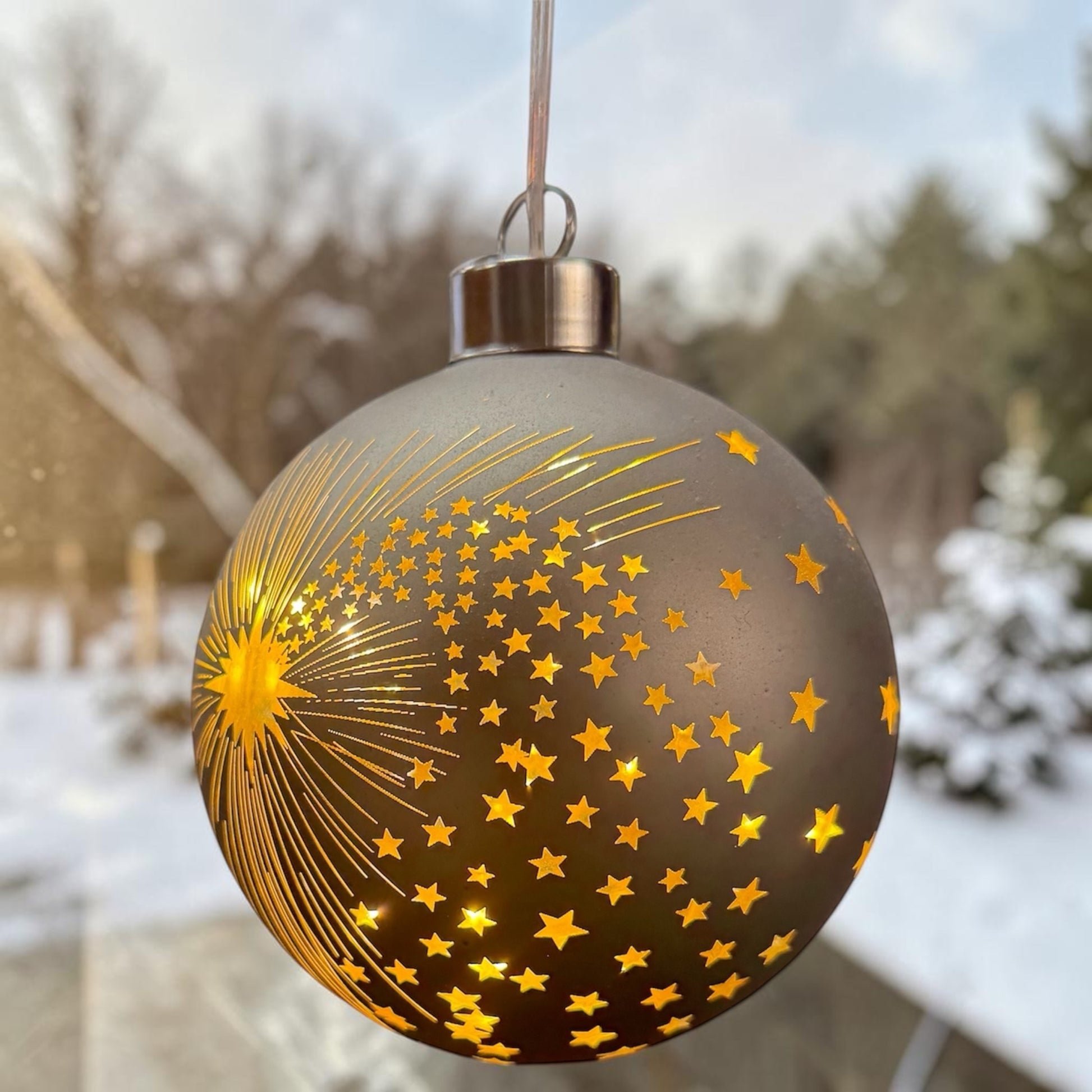 Eine taupefarbene LED-Dekokugel mit Sternschnuppenmotiv hängt vor einer Fensterfläche mit unscharfem Außenbereich.