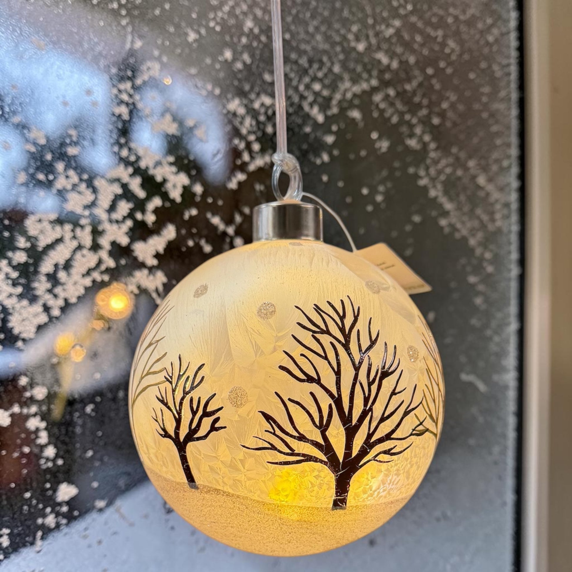 Eine matte, weiße LED-Dekokugel mit Baum-Motiv ist vor einem Fenster mit Schnee im Hintergrund positioniert.