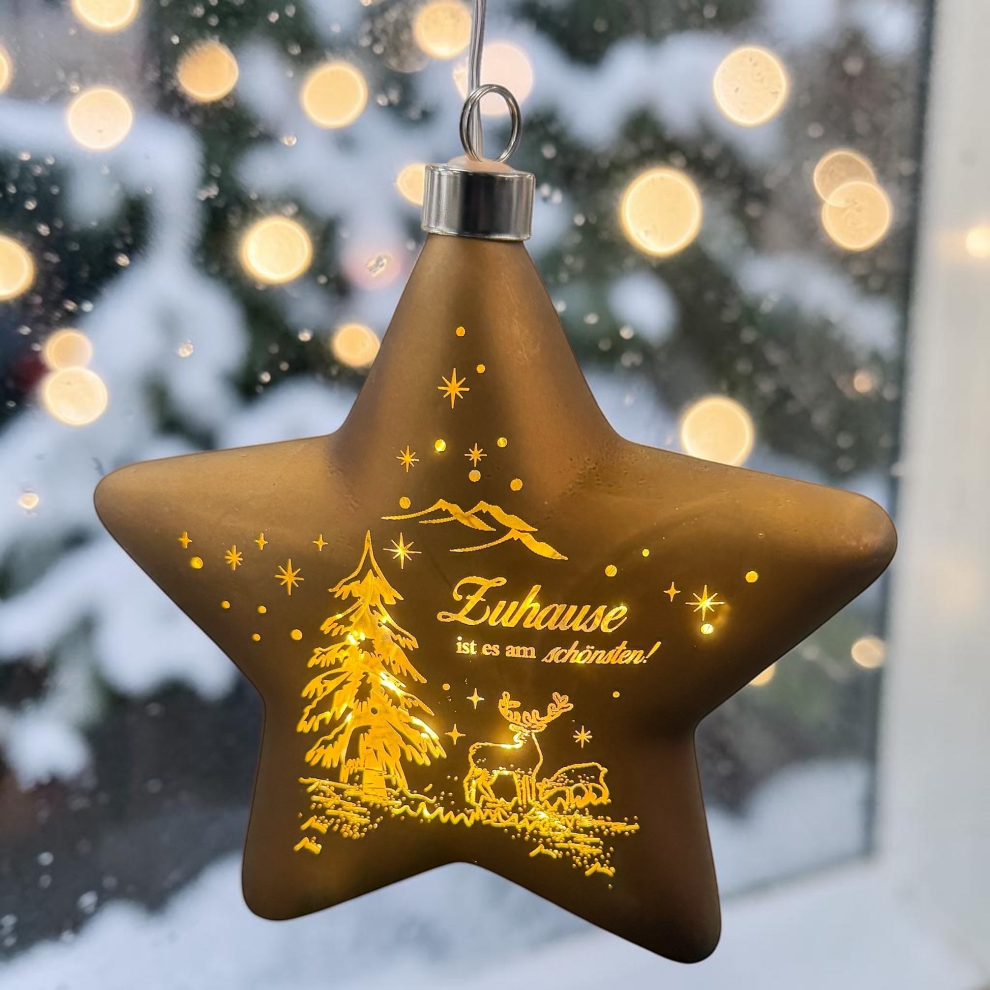 Ein sternförmiger, brauner LED-Dekoanhänger mit warmweißem Licht zeigt eine winterliche Szene mit Tannenbaum, Hirsch und dem Schriftzug „Zuhause ist es am schönsten!“ vor verschneitem Hintergrund.