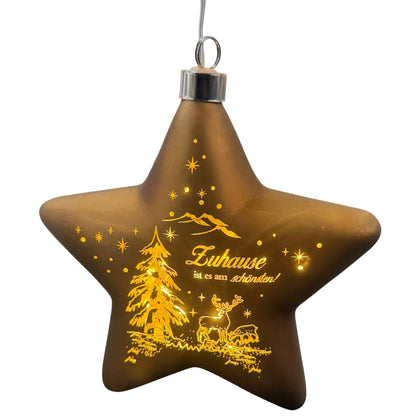 Ein sternförmiger, brauner LED-Dekoanhänger mit warmweißem Licht zeigt eine winterliche Szene mit Tannenbaum, Hirsch und dem Schriftzug „Zuhause ist es am schönsten!“.