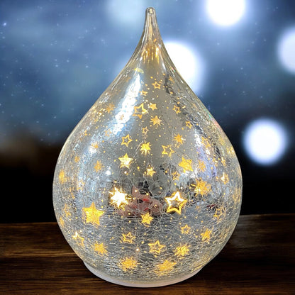 Ein silberner LED-Deko-Tropfen aus Crackle-Glas mit goldenen Sternen, strahlend im warmen Licht eines festlichen Ambientes.