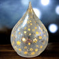 Ein silberner LED-Deko-Tropfen aus Crackle-Glas mit goldenen Sternen, strahlend im warmen Licht eines festlichen Ambientes.