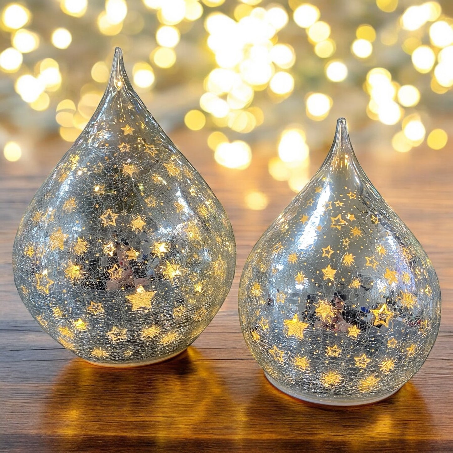 Leuchtende silberne Crackle-Glas-Tropfen mit goldenen Sternen auf Holztisch vor funkelndem Lichterhintergrund.