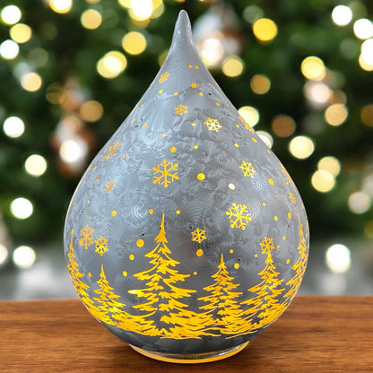 Silberner LED-Deko-Tropfen mit goldenen Tannen und Schneeflocken, strahlend vor weihnachtlichem Lichterhintergrund.