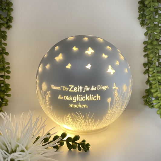 Eine silberne LED Dekolicht Kugel mit dem Spruch "Nimm' Dir Zeit für die Dinge, die Dich glücklich machen." und kleinen Schmetterlingen ist 15cm groß und beleuchtet im Ambiente platziert.