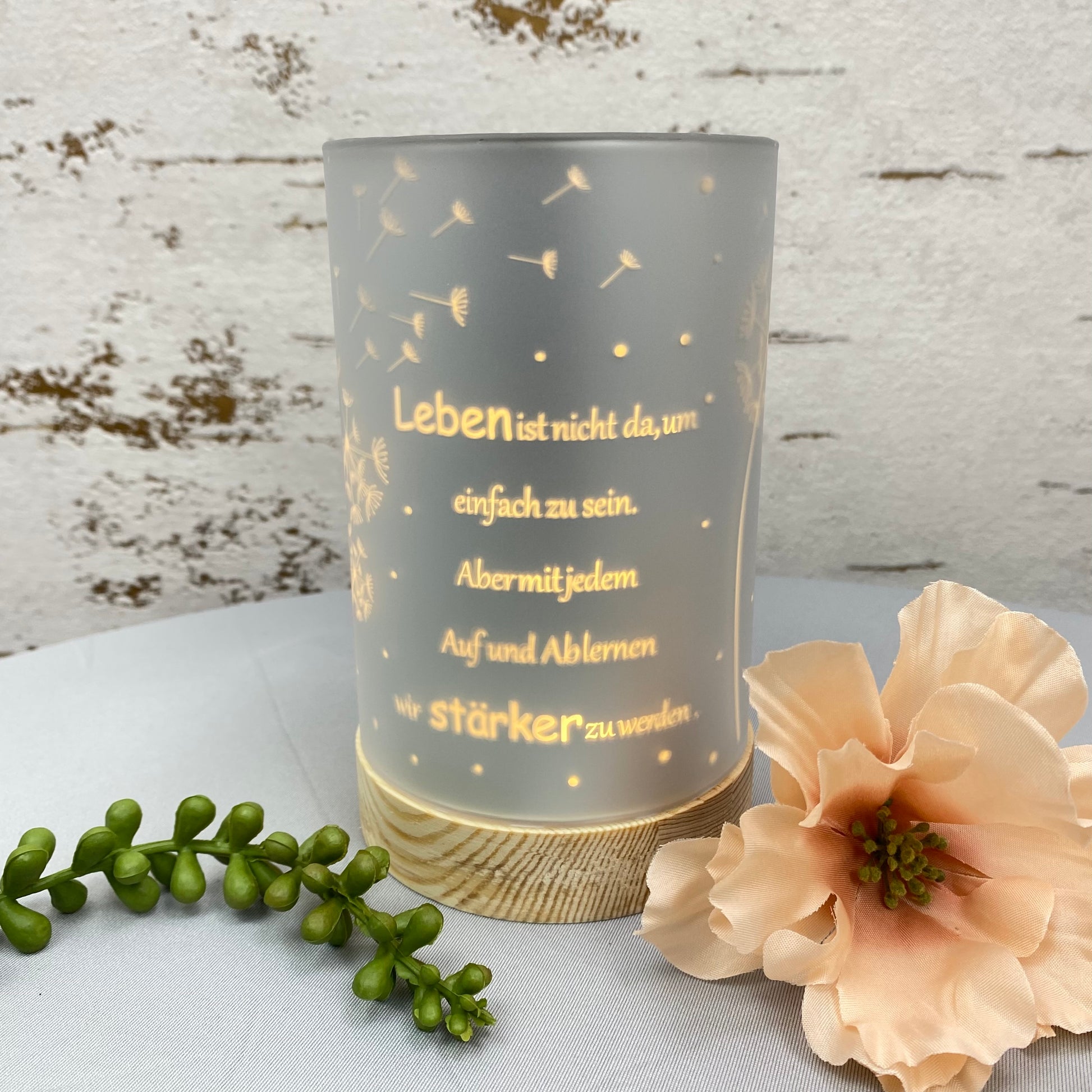 Eine silberne LED Säule mit Holz Sockel. Auf dem Dekolicht ist der Spruch "Leben ist nicht da, um einfach zu sein. Aber mit jedem Auf und Ab lernen wir stärker zu werden." und Pusteblumen abgebildet in goldener Schrift. Das Dekolicht ist 15cm groß und beleuchtet im Ambiente platziert.