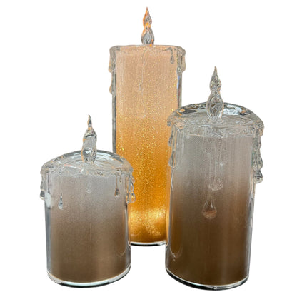 Drei LED-Kerzen aus transparentem Acryl in unterschiedlichen Größen. Zylindrische Form mit angedeuteten Wachstropfen am oberen Rand und flammenförmiger Spitze. Innen goldfarbene Struktur.