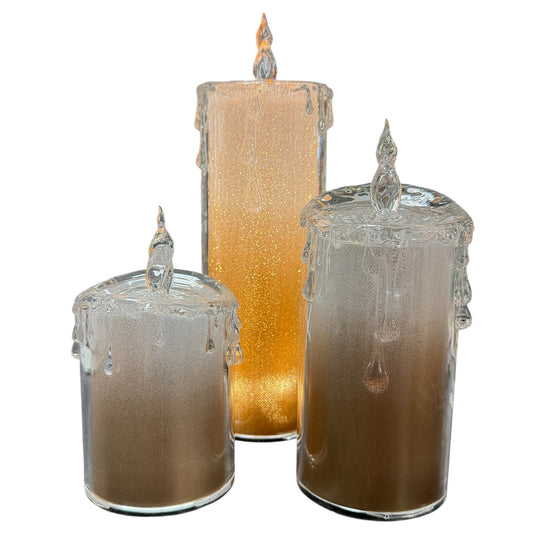 Drei LED-Kerzen aus transparentem Acryl in unterschiedlichen Größen. Zylindrische Form mit angedeuteten Wachstropfen am oberen Rand und flammenförmiger Spitze. Innen goldfarbene Struktur.