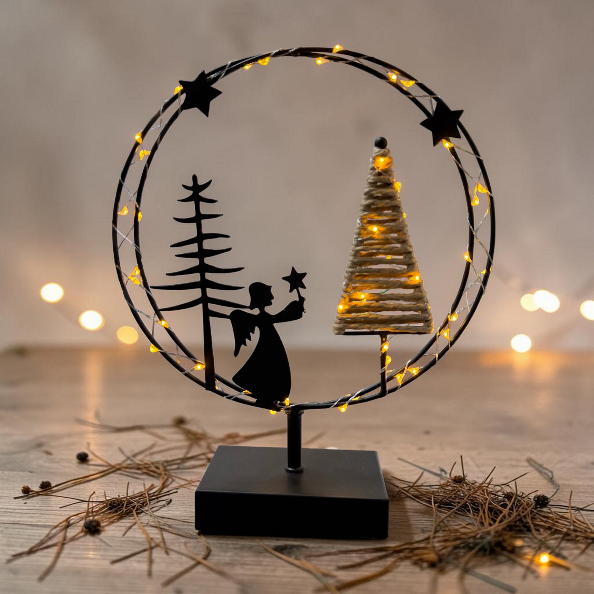 Ein runder Metallkranz mit integrierter warmweißer LED-Lichterkette steht auf einem schwarzen Sockel und zeigt eine Szene mit Engel, Tannenbaum und Stern vor stimmungsvoll beleuchtetem Hintergrund.