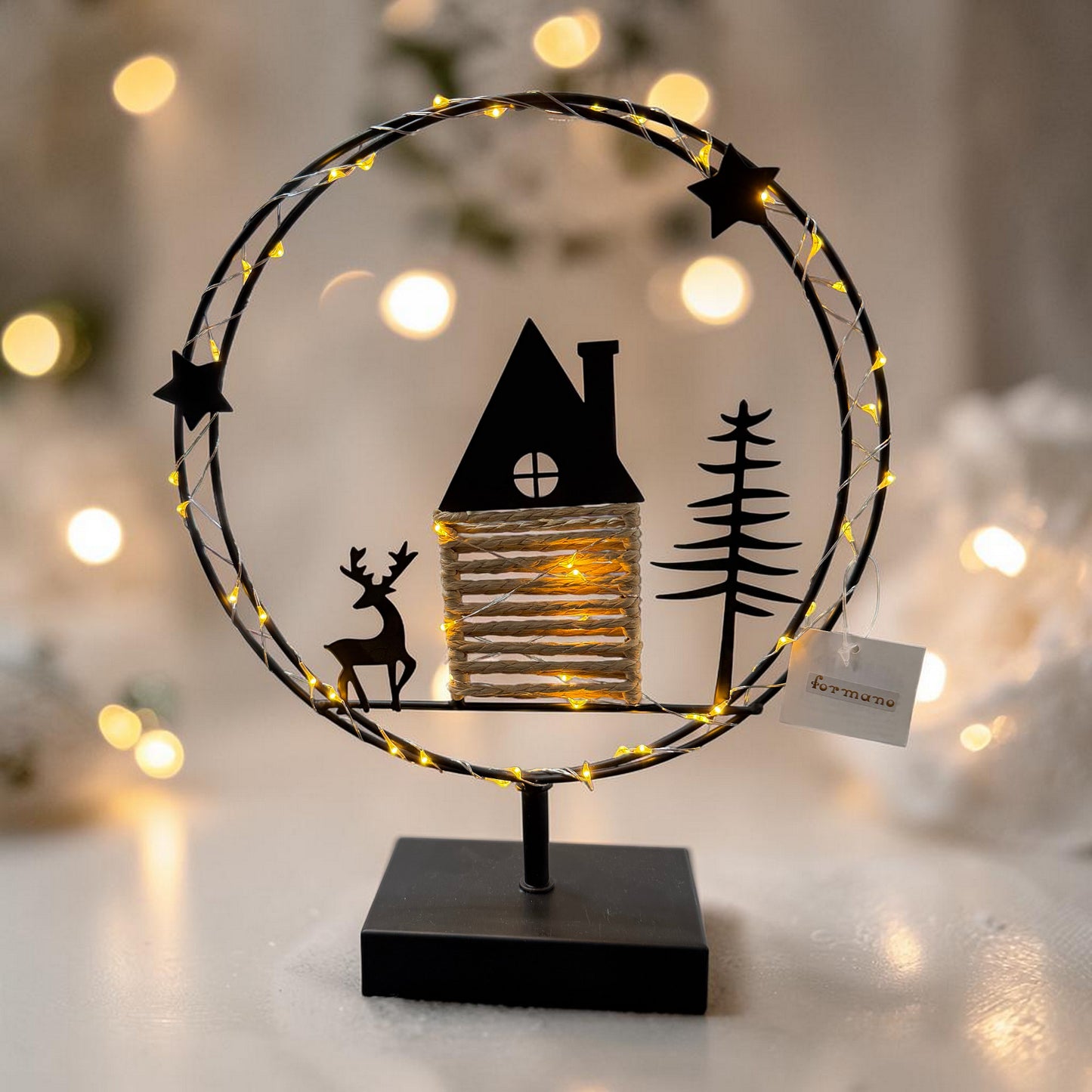 Ein runder Metallkranz mit integrierter warmweißer LED-Lichterkette steht auf einem schwarzen Sockel und zeigt eine Szene mit Haus, Hirsch und Tannenbaum vor stimmungsvoll beleuchtetem Hintergrund.