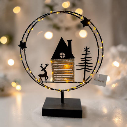 Ein runder Metallkranz mit integrierter warmweißer LED-Lichterkette steht auf einem schwarzen Sockel und zeigt eine Szene mit Haus, Hirsch und Tannenbaum vor stimmungsvoll beleuchtetem Hintergrund.