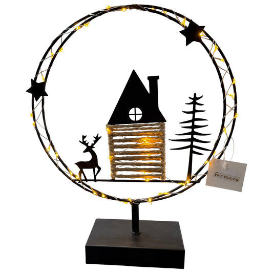 Ein runder Metallkranz mit integrierter warmweißer LED-Lichterkette steht auf einem schwarzen Sockel und zeigt eine Szene mit Haus, Hirsch und Tannenbaum.