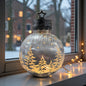 Eine silberne LED-Glaskugel mit goldener Winterlandschaft steht auf einer Fensterbank und leuchtet stimmungsvoll vor verschneiter Kulisse.